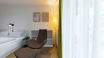 A modern hotel room with a bed, two brown chairs, a white nightstand, a floor lamp, and a bookshelf.