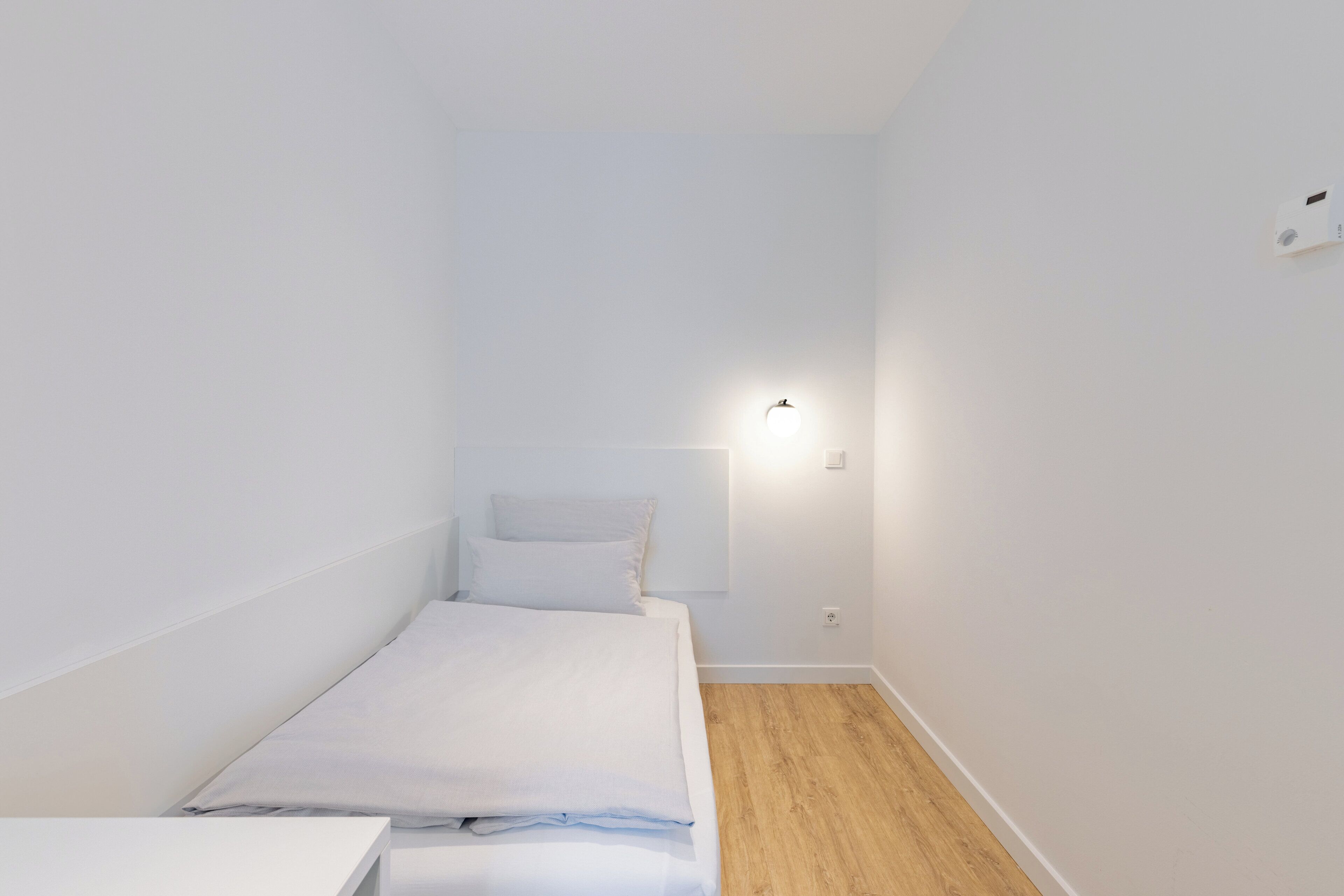 Een kleine, witte kamer met een enkel bed, houten vloer en een wandlamp.