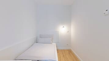 A small, white, single-bed room with a wooden floor and a wall-mounted light fixture.