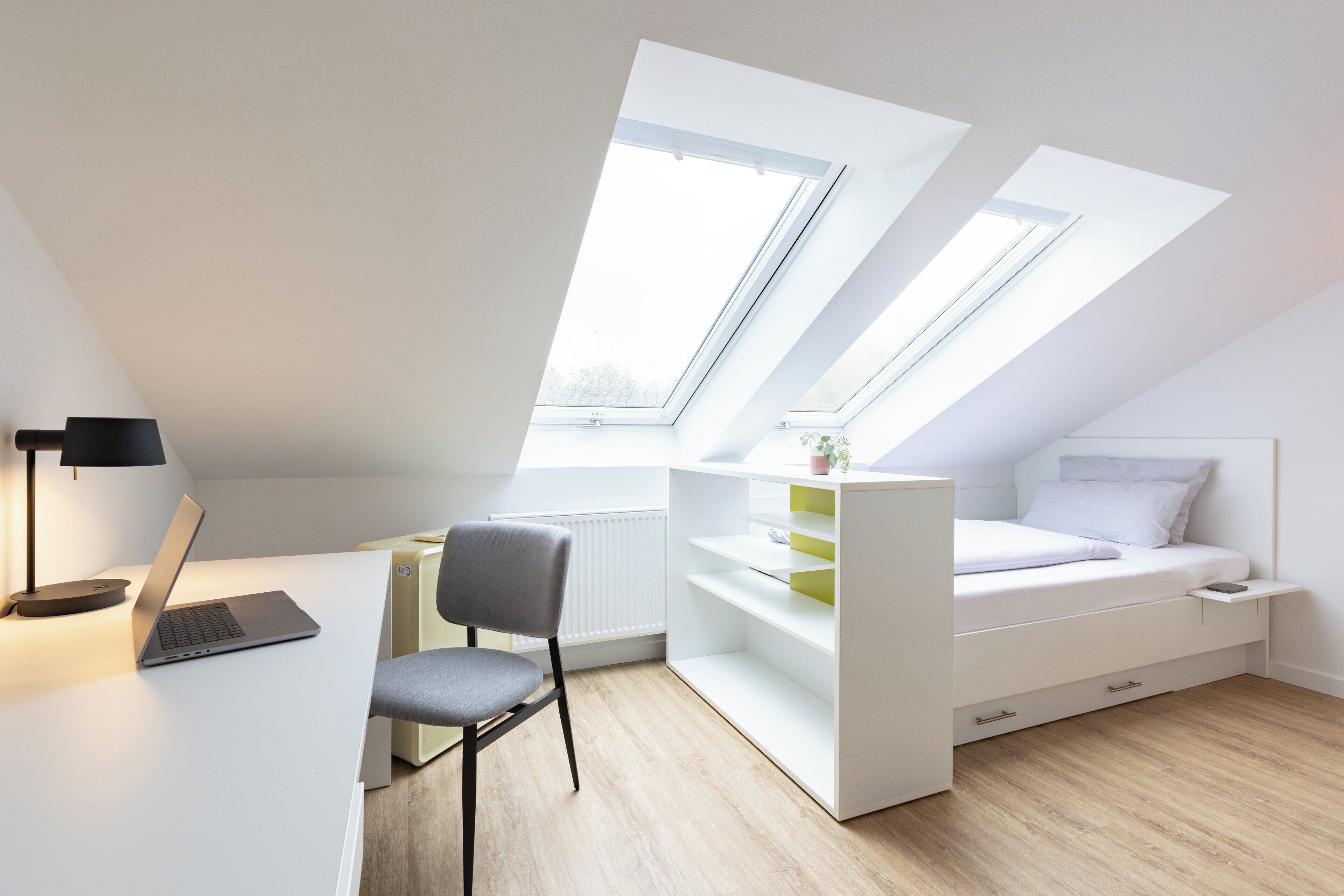Quarto moderno e compacto com claraboia, uma escrivaninha com um laptop e uma cama com gavetas embutidas.
