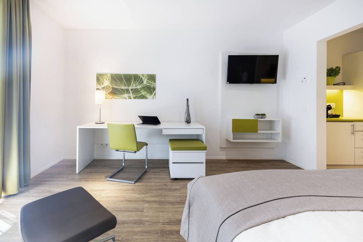 A modern hotel room with a bed, desk, chair, TV, and a small kitchenette.
