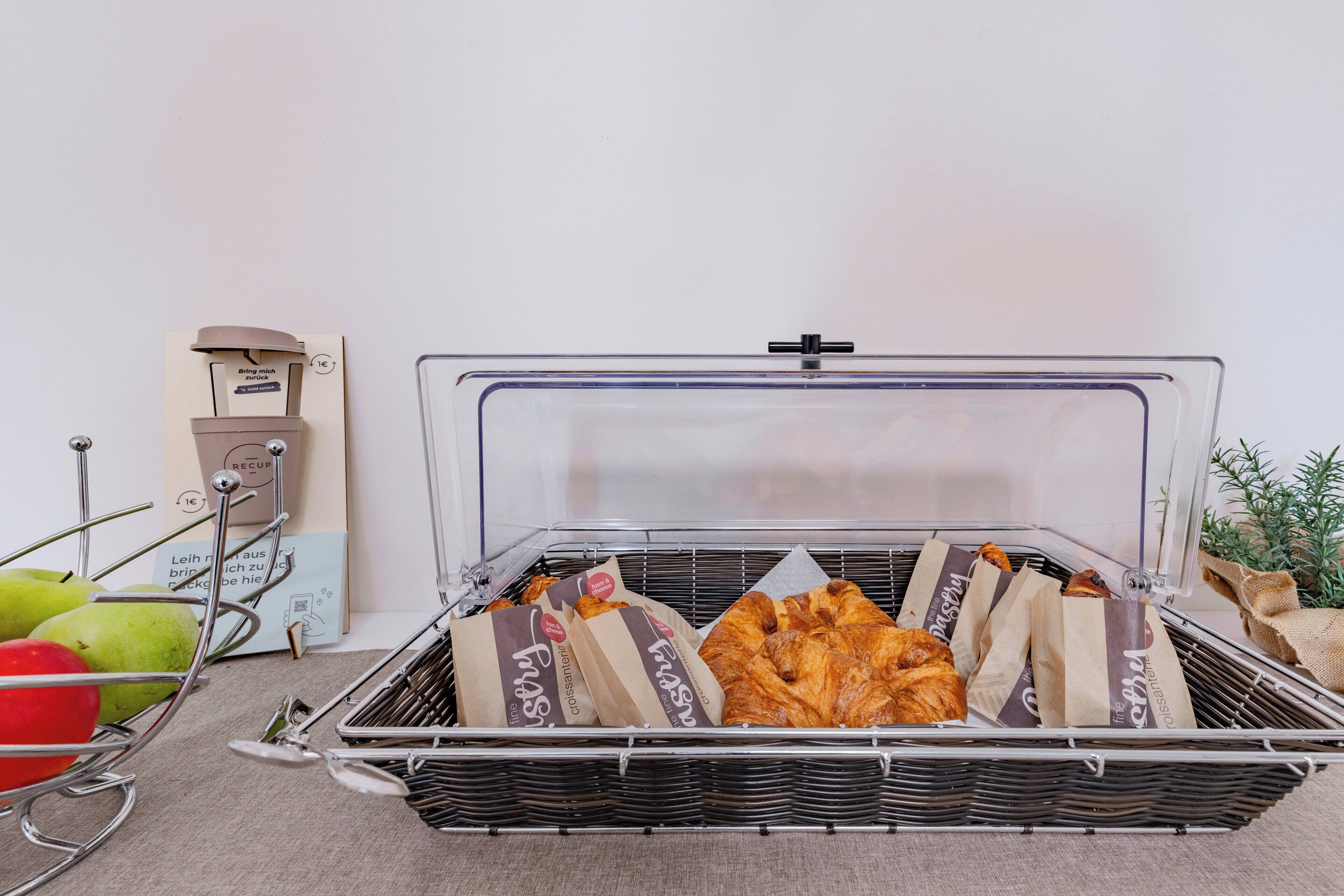 A wicker basket with a clear lid containing pastries and a baguette, placed on a carpeted floor next to a metal rack with apples and a coffee machine.