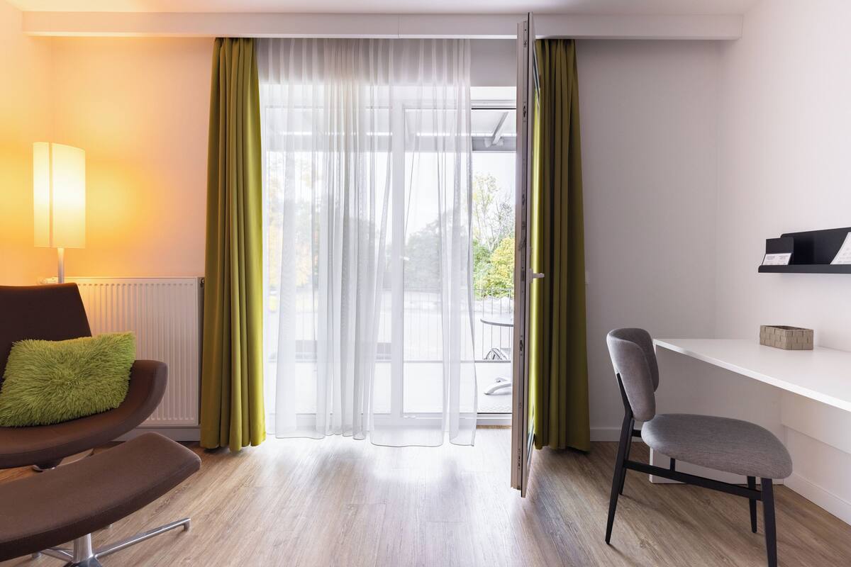 A modern room with a desk, a chair, a lamp, and a view through a large window with sheer curtains.