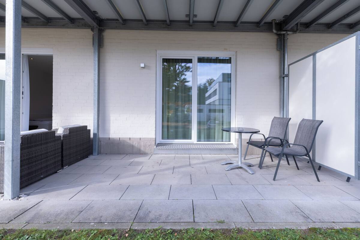 An outdoor patio with a table and chairs, a sliding glass door, and a covered area with a white wall and metal beams.
