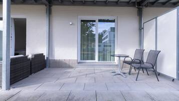 Un patio exterior con mesa y sillas, un ventanal corredero y una zona cubierta con pared blanca y vigas metálicas.