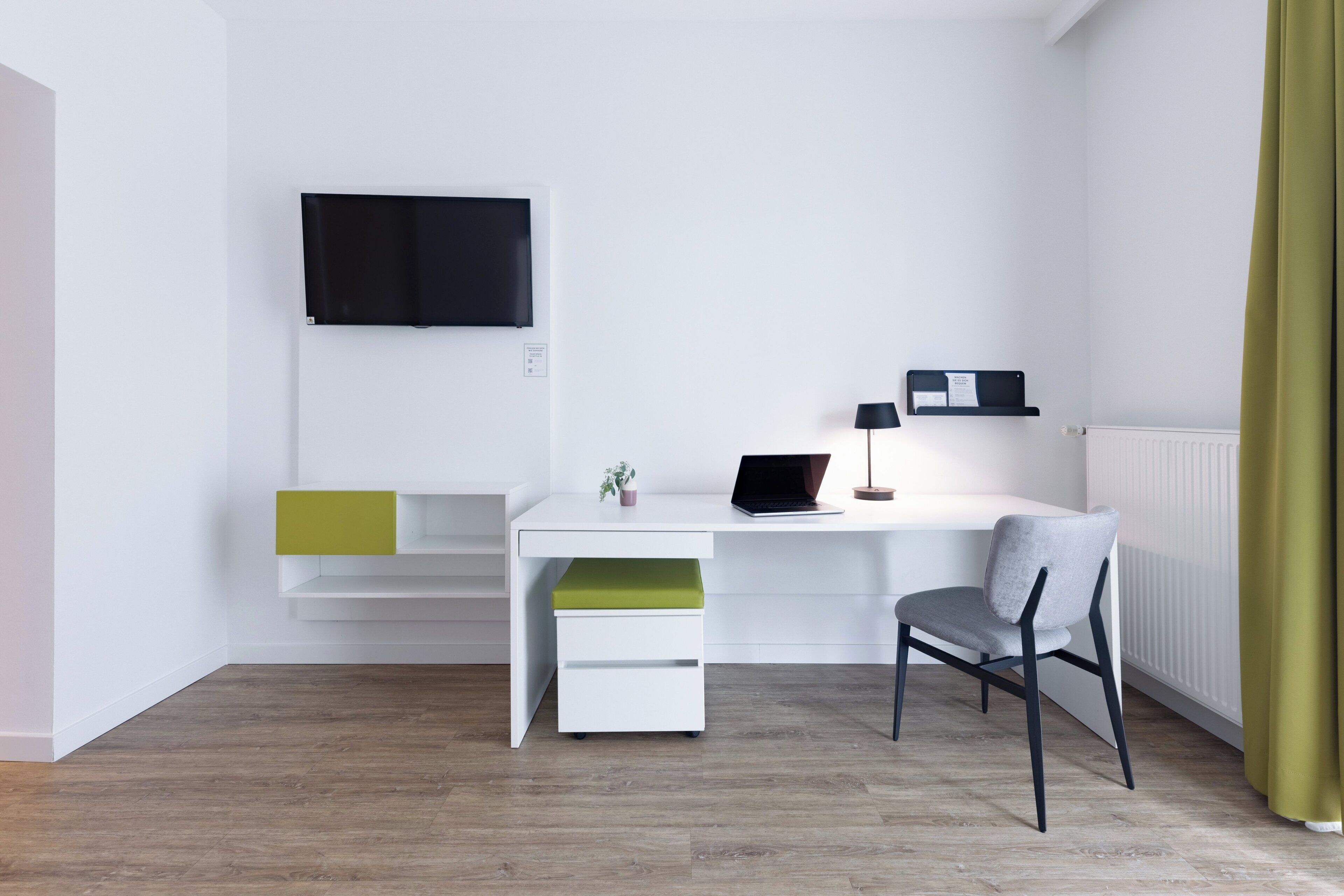 A modern, minimalist room with a white desk, a laptop, a flat-screen TV, and a chair.