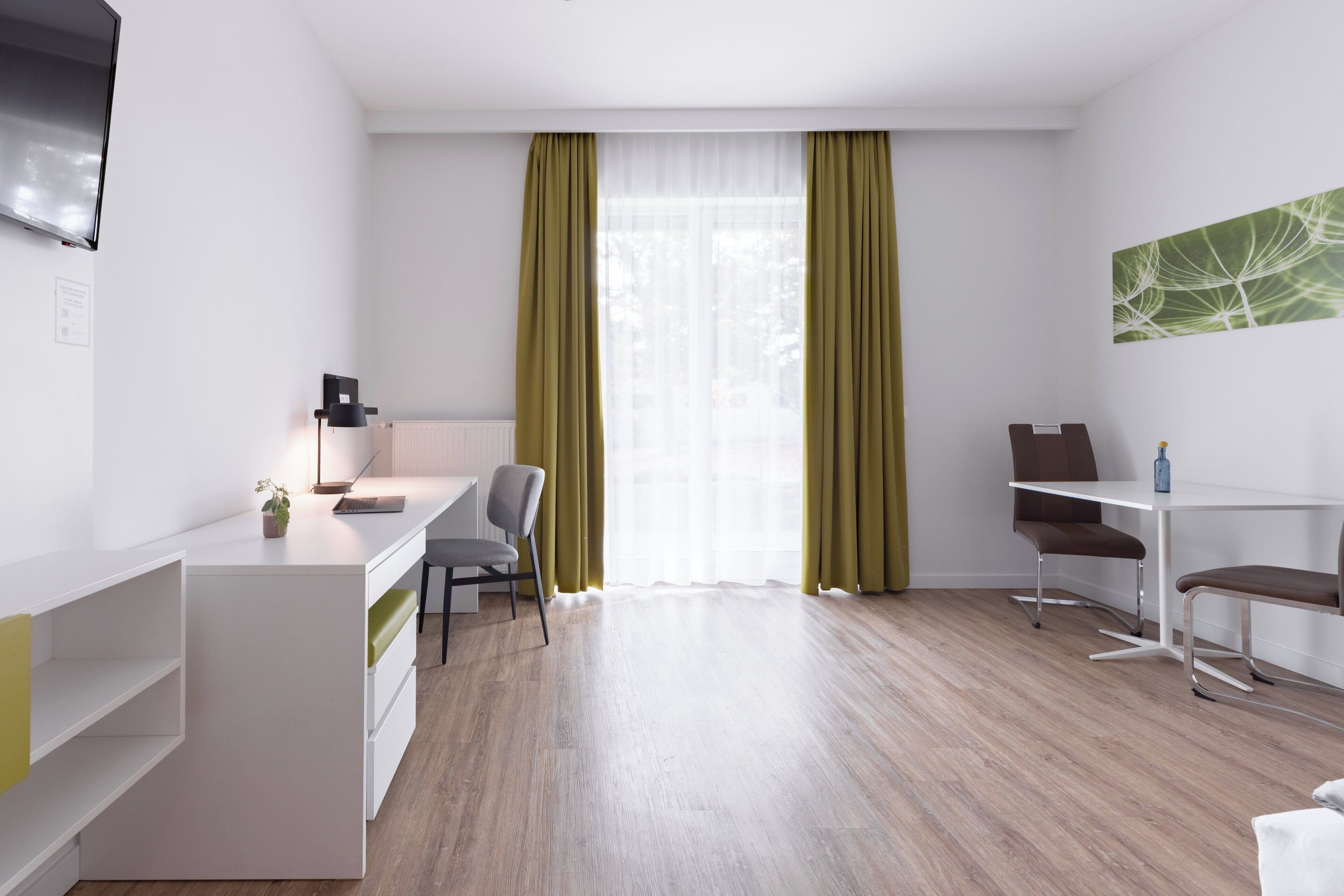 A modern, well-lit room with a desk, two chairs, a small table, and a large window with sheer curtains.