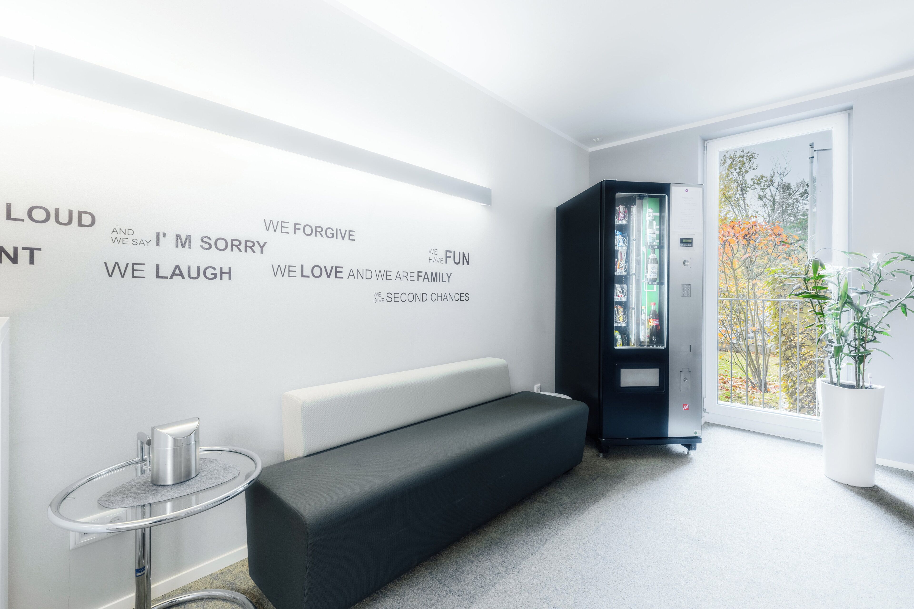 A modern room with a black sofa, a small table with a kettle, a bookshelf, and a large window with a view of trees.