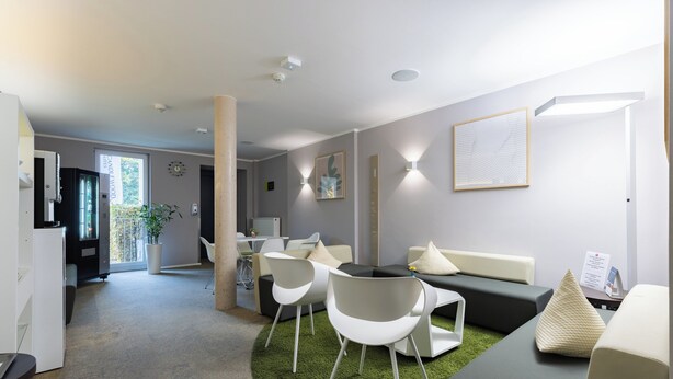 A modern, well-lit room with a green carpet, white chairs, a grey sofa, and a small table. - Candlewood Suites Furth by IHG (Fuerth)