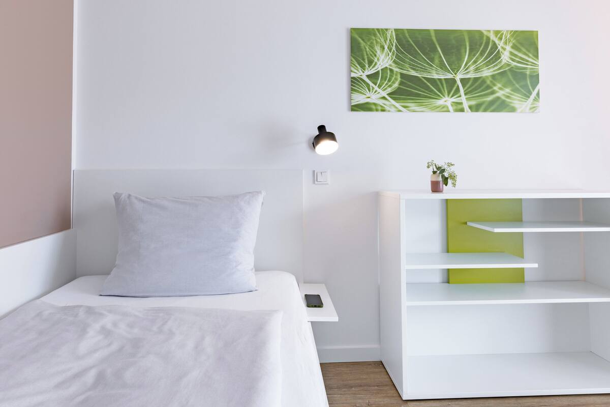 A modern bedroom with a white bed, a nightstand, a wall-mounted lamp, a green-framed artwork, and a white shelving unit.