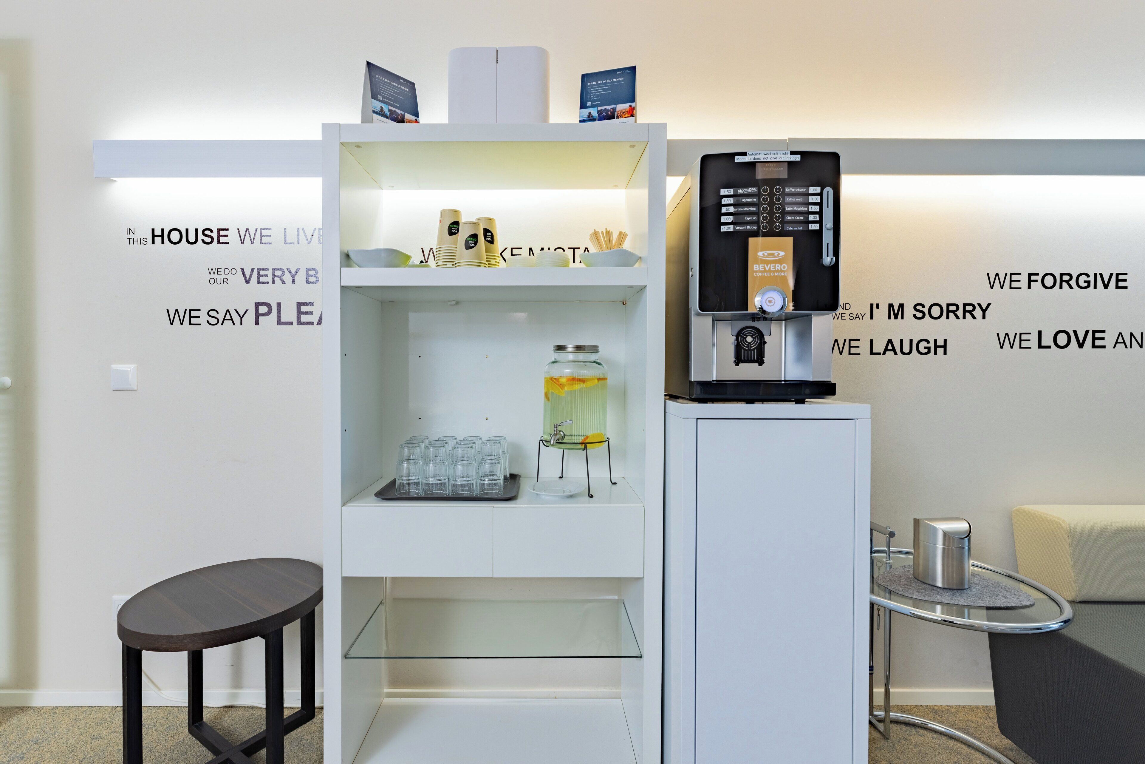 Een moderne hotel receptie met een wit balie, een koffieapparaat en een glazen vitrine met diverse artikelen.