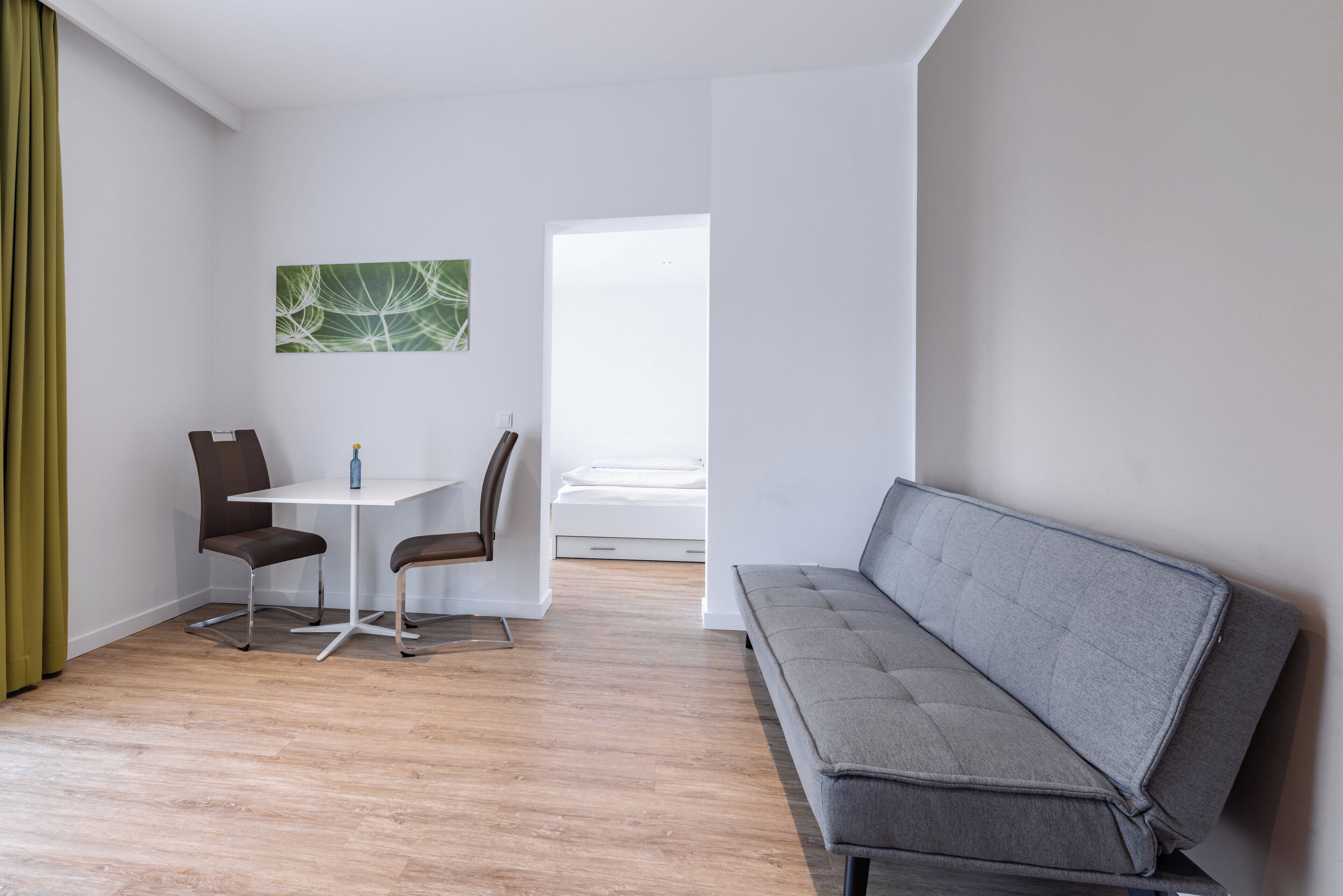 Un salon moderne avec un canapé gris, une petite table blanche, deux fauteuils marron et un rideau vert.