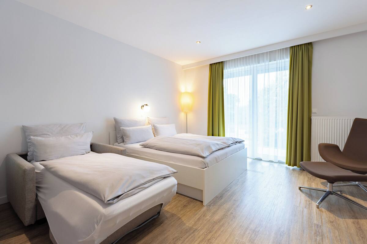 A hotel room with two beds, a chair, a lamp, and a window with curtains.