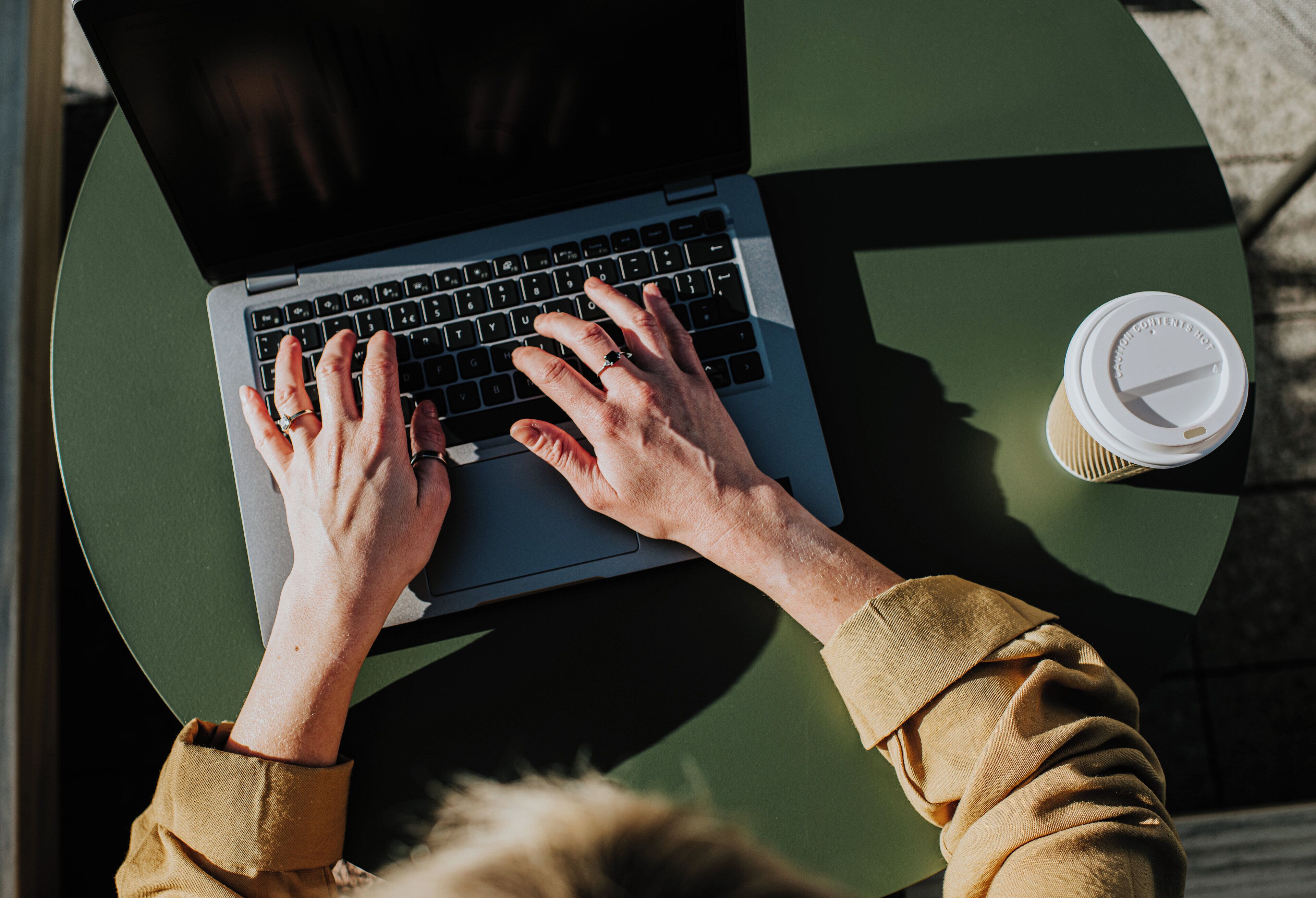 A person using a laptop on a green table with a coffee cup beside it.