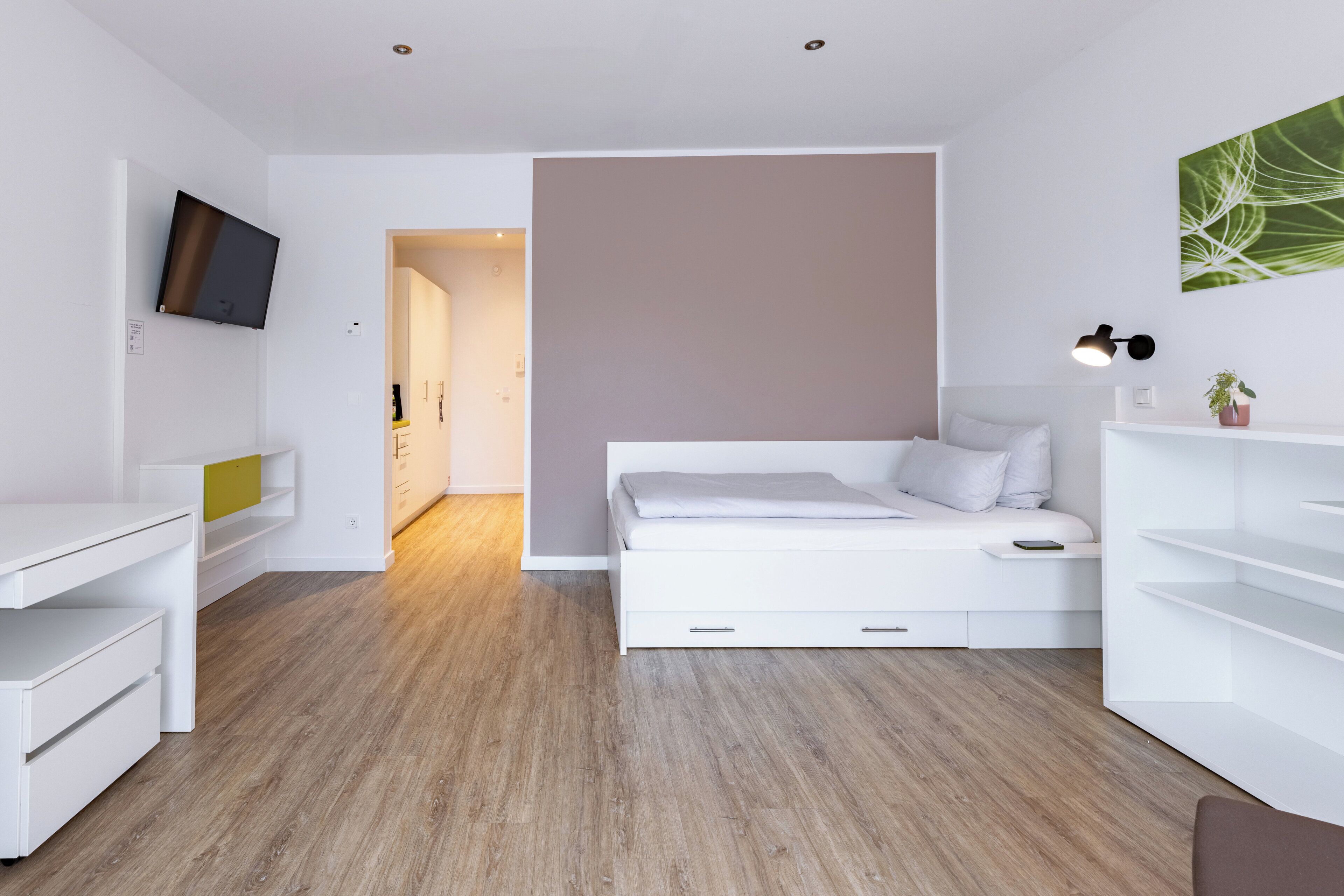 A modern, minimalist bedroom with a white bed, built-in desk, and a TV mounted on the wall.