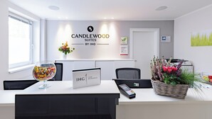 A modern reception desk with a sign for Candlewood Suites by IHG, a glass vase with flowers, a basket of flowers, and a remote control. - Candlewood Suites Furth by IHG (Fuerth)