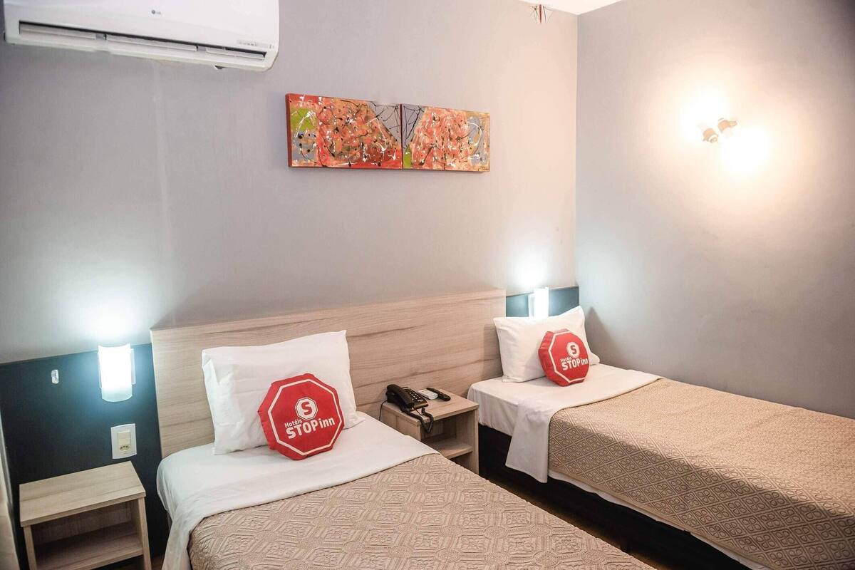 Quarto de hotel com duas camas, mesa de cabeceira com telefone, ar condicionado fixado na parede e um quadro sobre as camas.