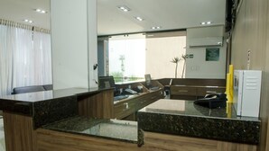 Zona de recepción con mostrador de madera, computadora y teléfono. Se ve una ventana con cortinas y una planta al fondo.