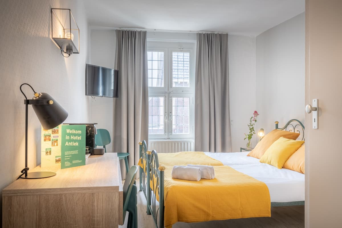 A hotel room with a bed, a desk with a lamp, a TV, and a window with curtains.