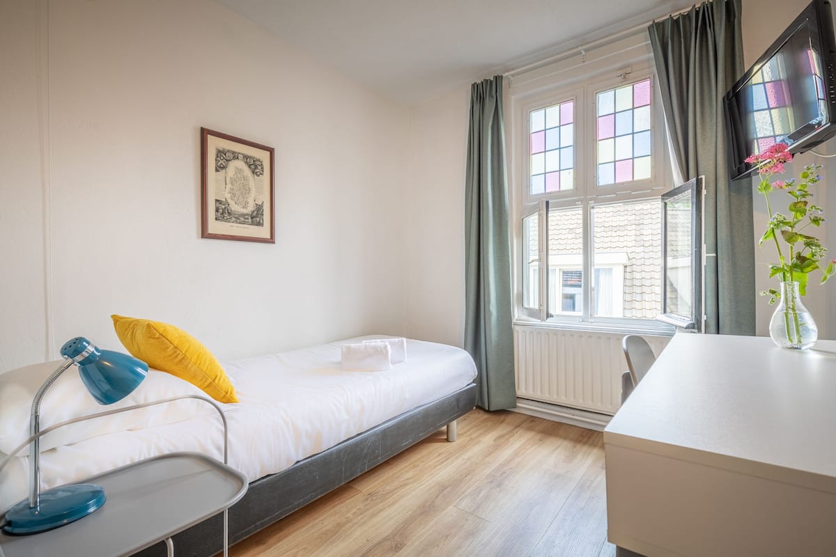 A hotel room with a single bed, a desk with a vase of flowers, a picture on the wall, and a window with curtains.