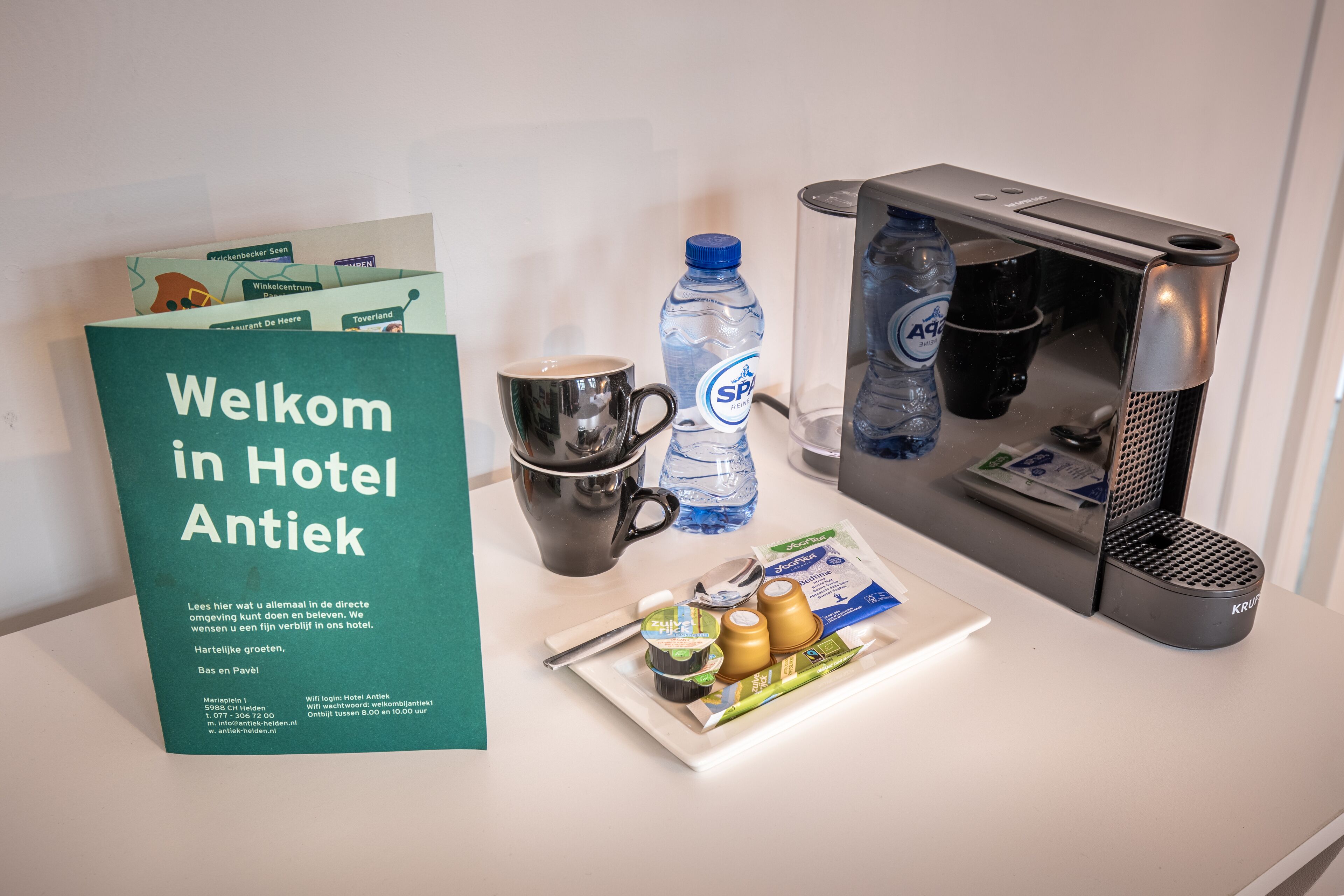 Habitación de hotel con un cartel de bienvenida, una taza de café, una botella de agua, una tetera y una bandeja con condimentos.