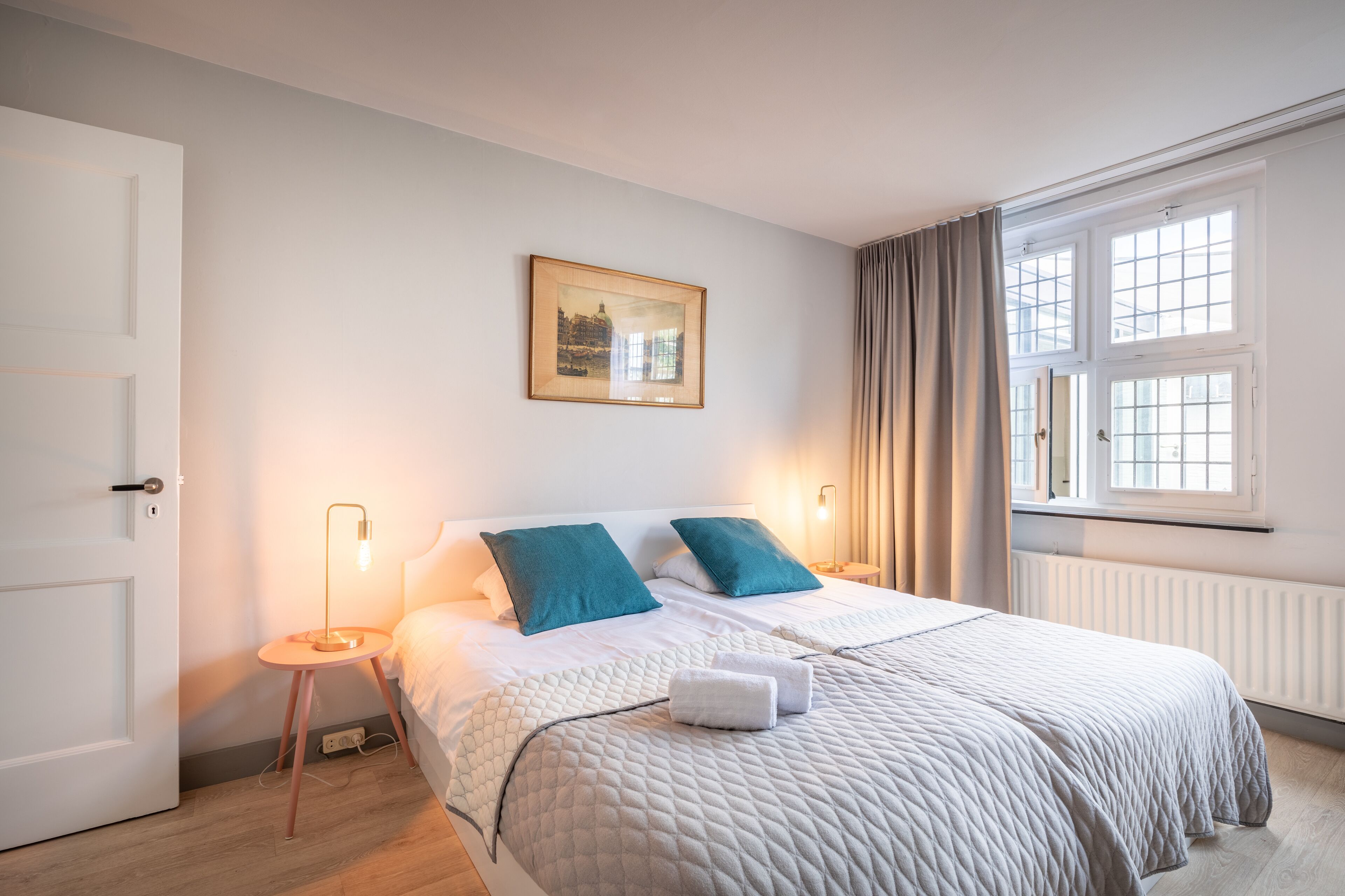 Una camera da letto ordinata con un letto, abat-jour, un quadro appeso e una finestra con tende.
