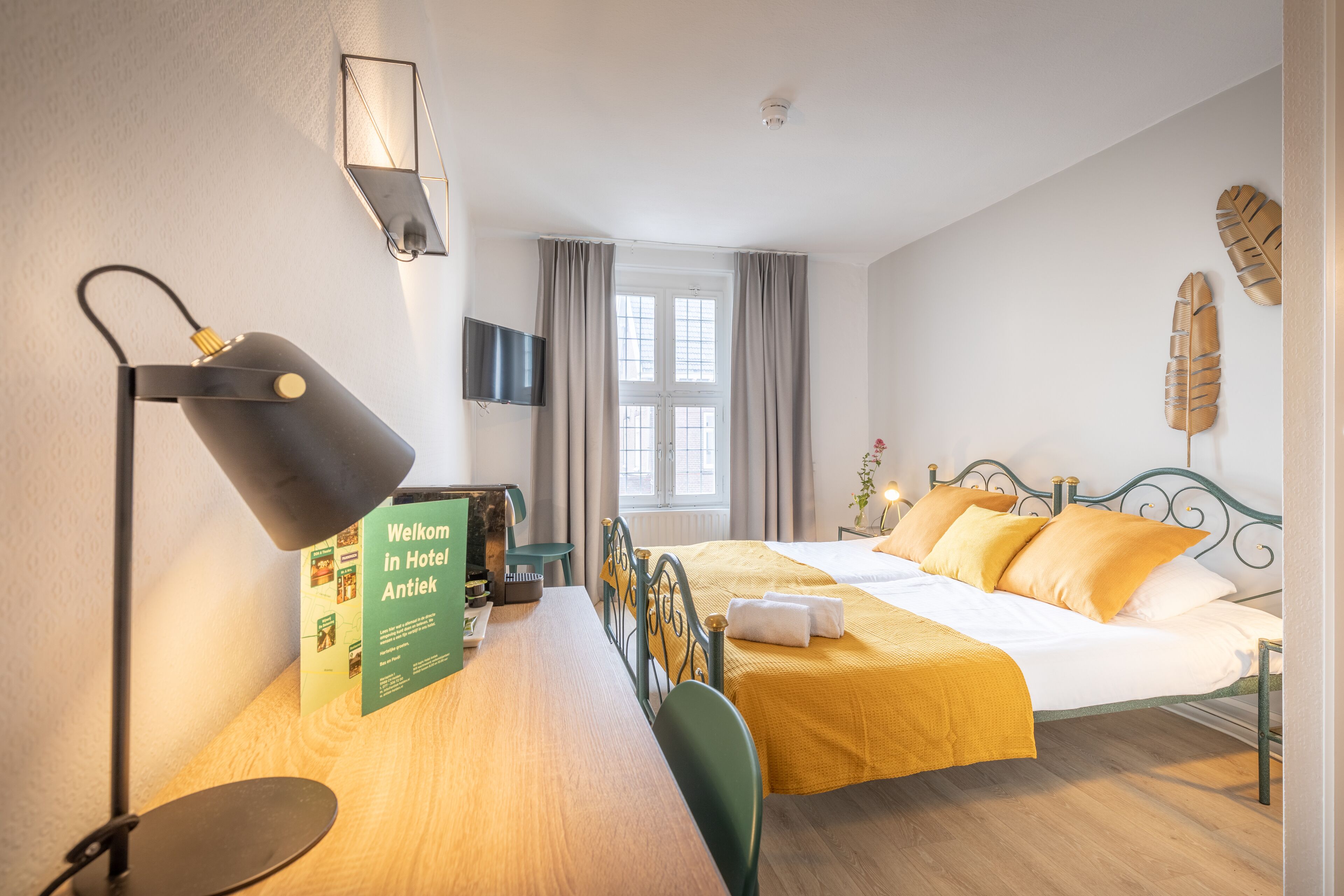 Une chambre d’hôtel avec un lit, un bureau avec une lampe, une chaise, une télévision et une fenêtre avec des rideaux.