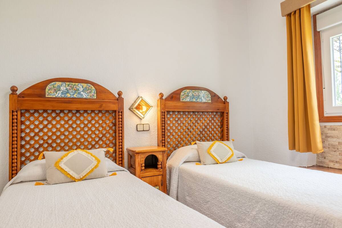 A hotel room with two beds, wooden headboards, a small wooden nightstand, and a window with curtains.