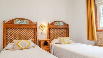 Quarto de hotel com duas camas, cabeceiras de madeira, uma mesinha de cabeceira de madeira e uma janela com cortinas.