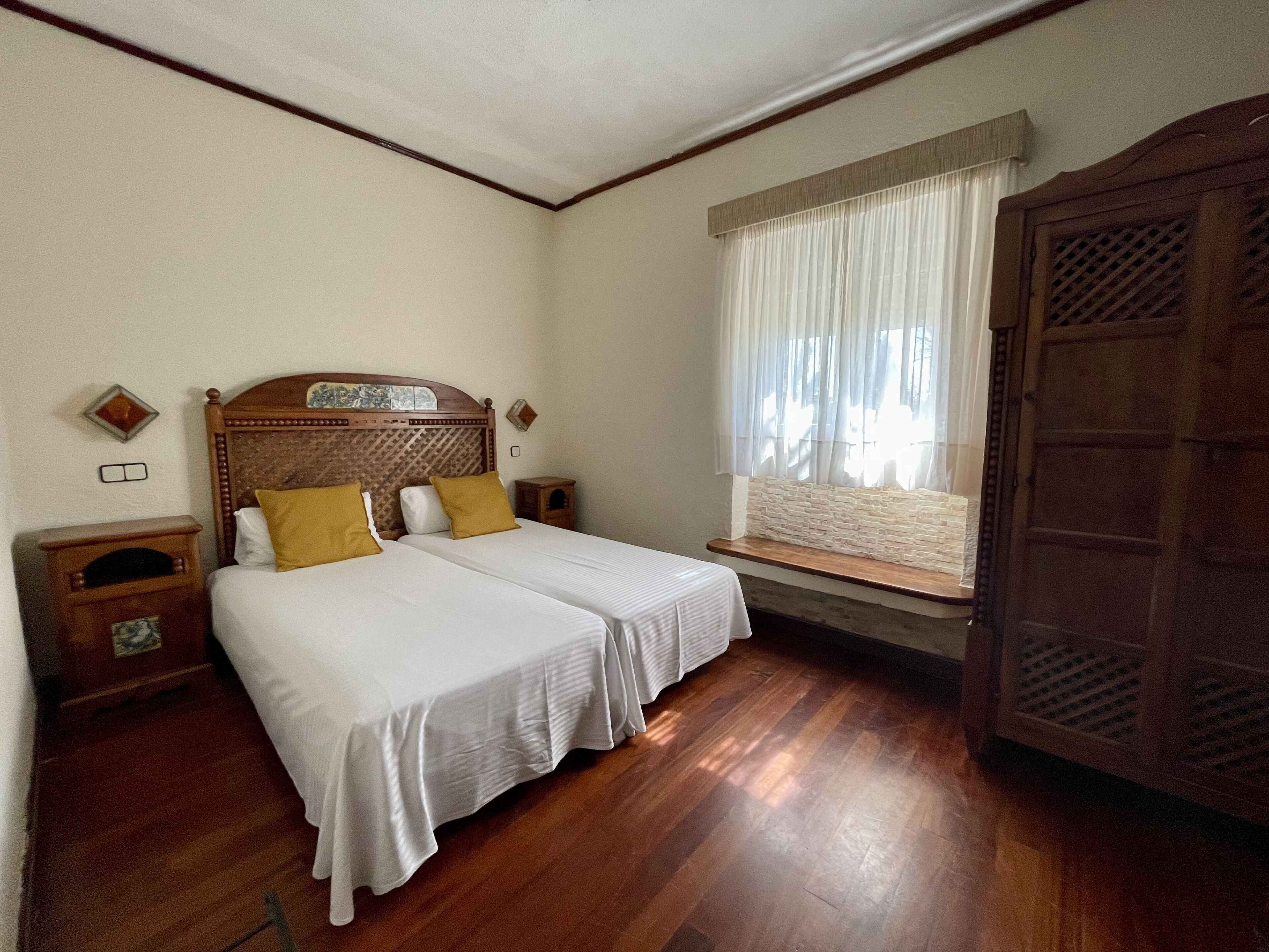 A room with two beds, wooden headboards, a window with curtains, a wooden wardrobe, and a nightstand with a lamp.
