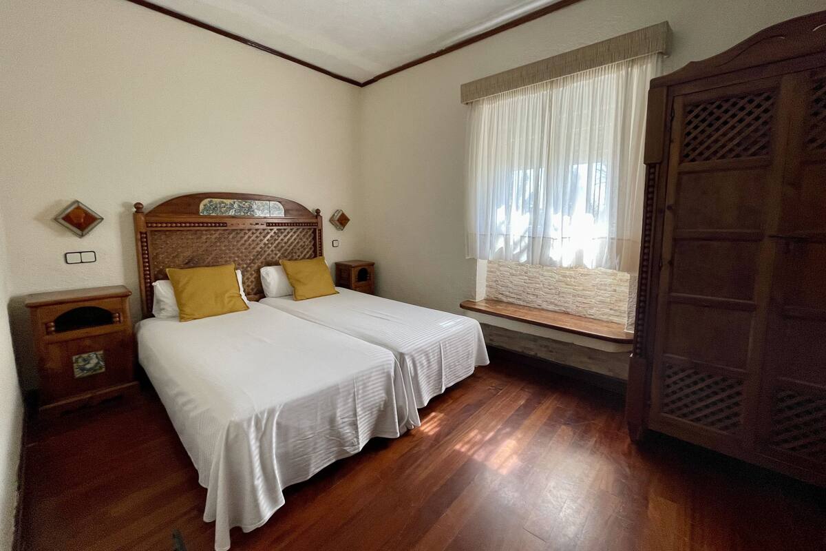 Una habitación con dos camas, cabeceros de madera, una ventana con cortinas, un armario de madera y una mesita de noche con lámpara.