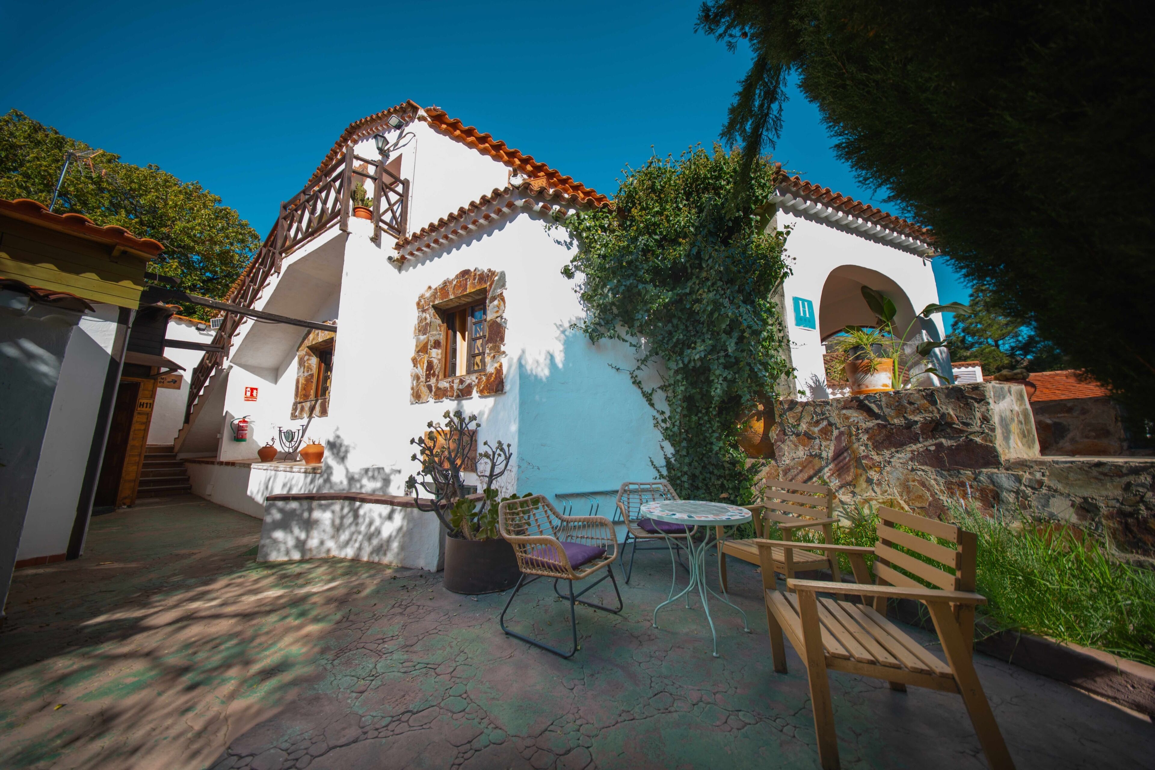 A white building with a tiled roof, a staircase, and a small patio area with a table, chairs, and a bench.