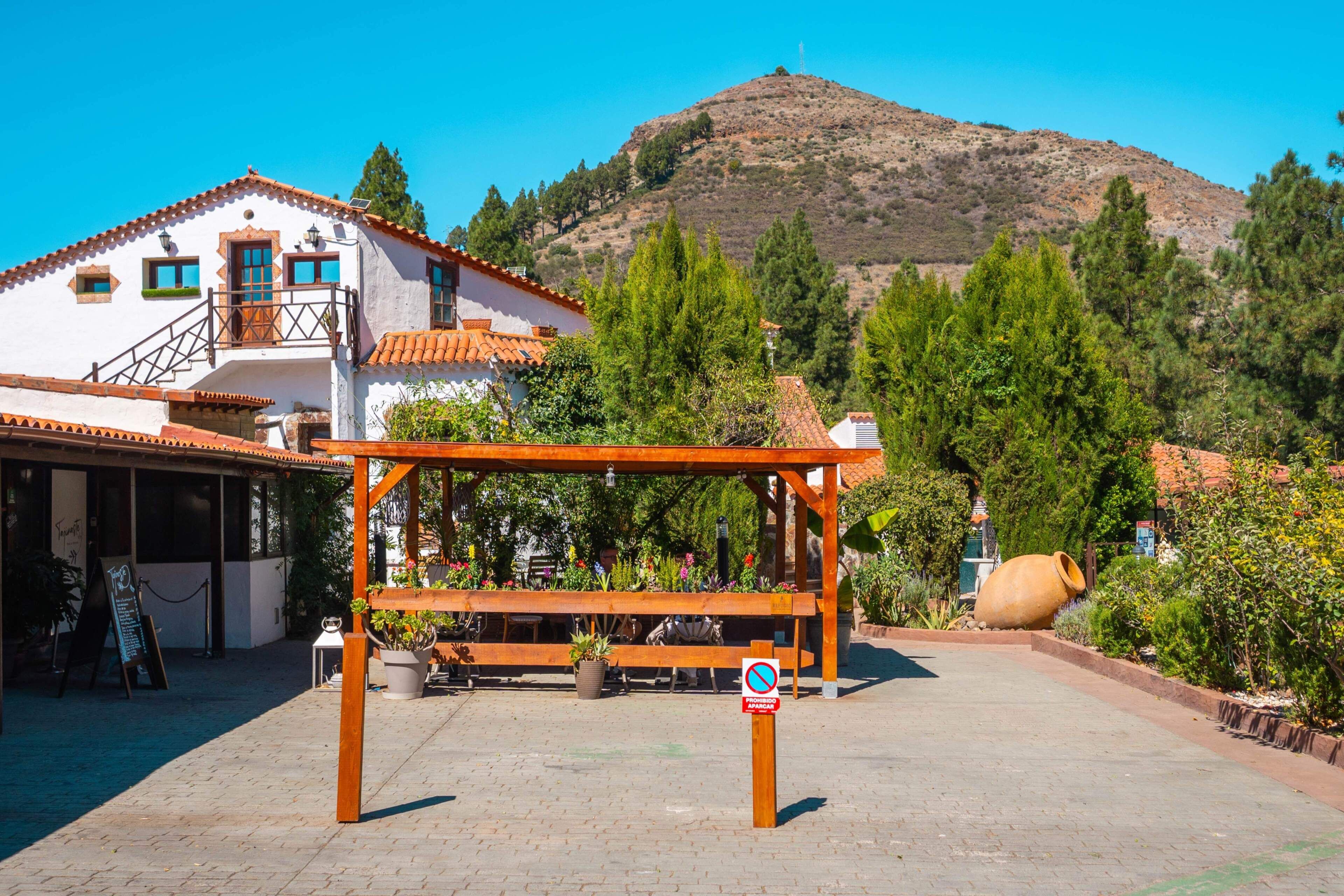 A courtyard with a wooden pergola, a large clay pot, and a mountain in the background.