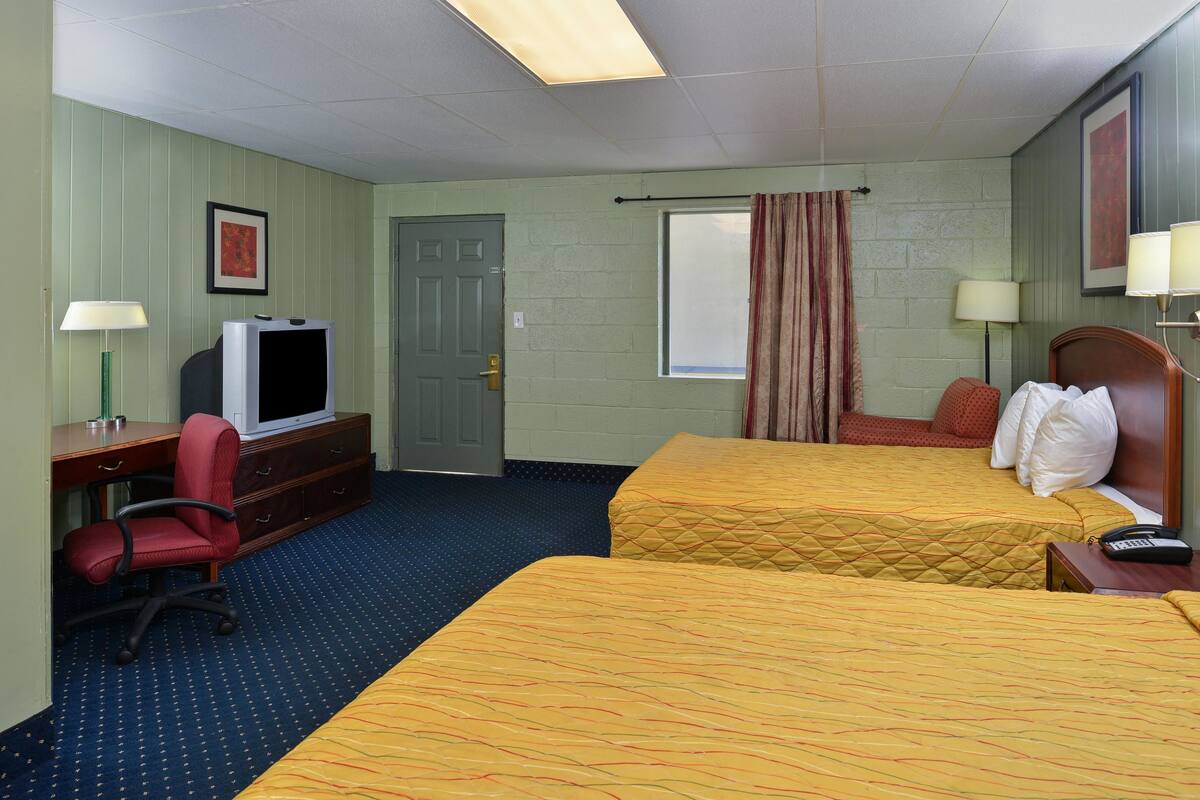 Ein Hotelzimmer mit zwei Betten, einem Schreibtisch mit Fernseher, einem Sessel und einem Fenster mit Vorhängen.