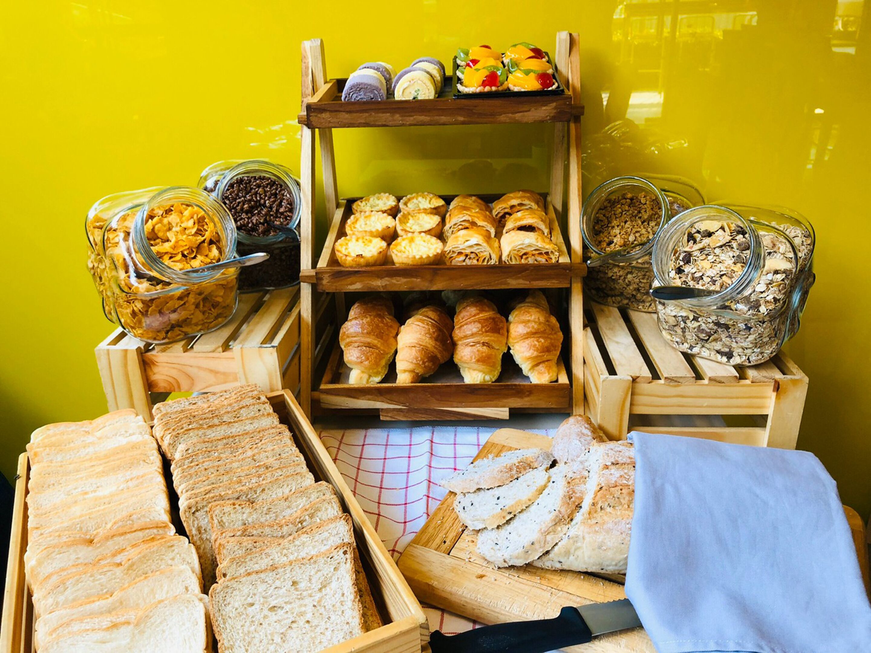 Ett urval av bakverk, inklusive bröd, croissanter, muffins och andra söta bakelser, som presenteras i trälådor och glasburkar.