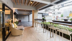An outdoor seating area with wicker chairs and a sofa, a bar counter, and a view of the street. - Alt Hotel Nana by UHG (Bangkok)