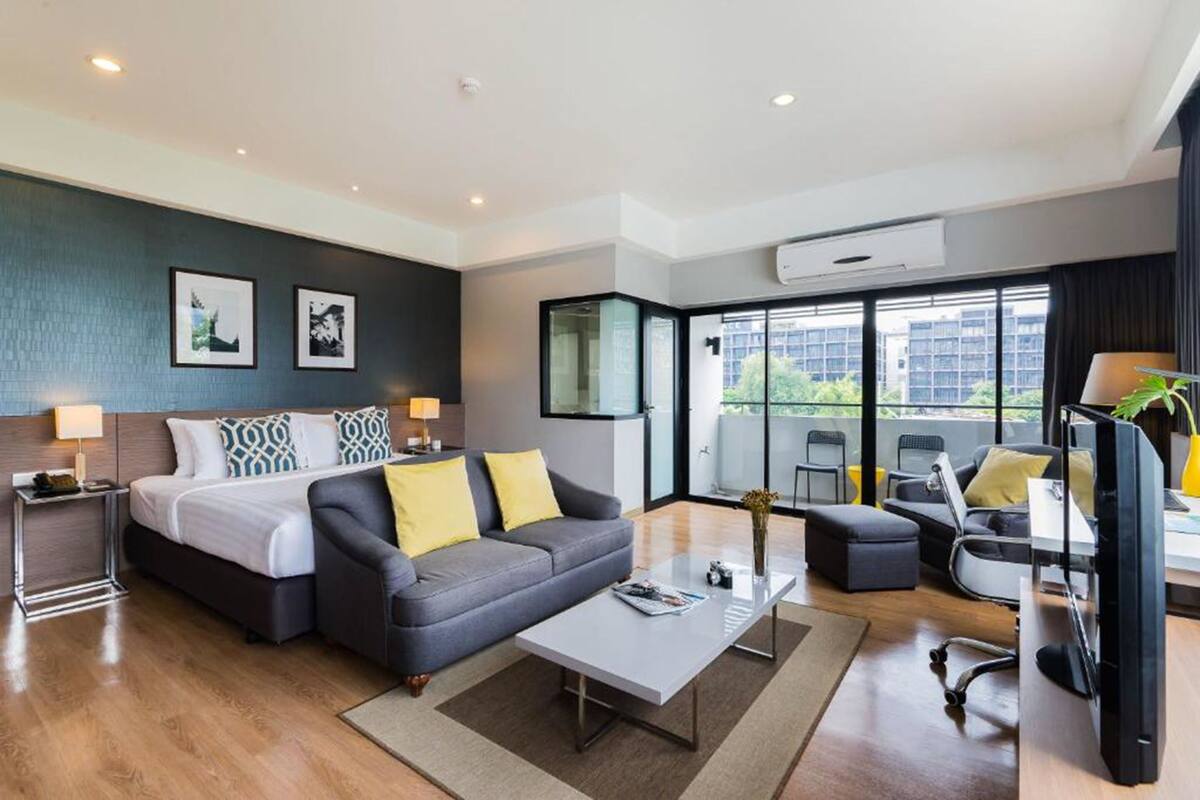 A modern hotel room with a bed, sofa, coffee table, and a desk with a computer. There are two framed pictures on the wall, a window with a view of buildings, and an air conditioning unit.