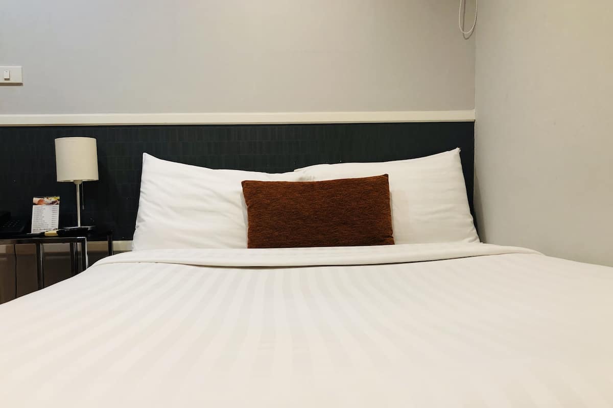 A neatly made bed with white bedding and a brown accent pillow, a bedside table with a lamp, and a wall-mounted phone.