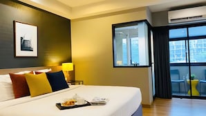 A hotel room with a large bed, a bedside table with a lamp, a framed picture on the wall, and a balcony with chairs. - Alt Hotel Nana by UHG (Bangkok)