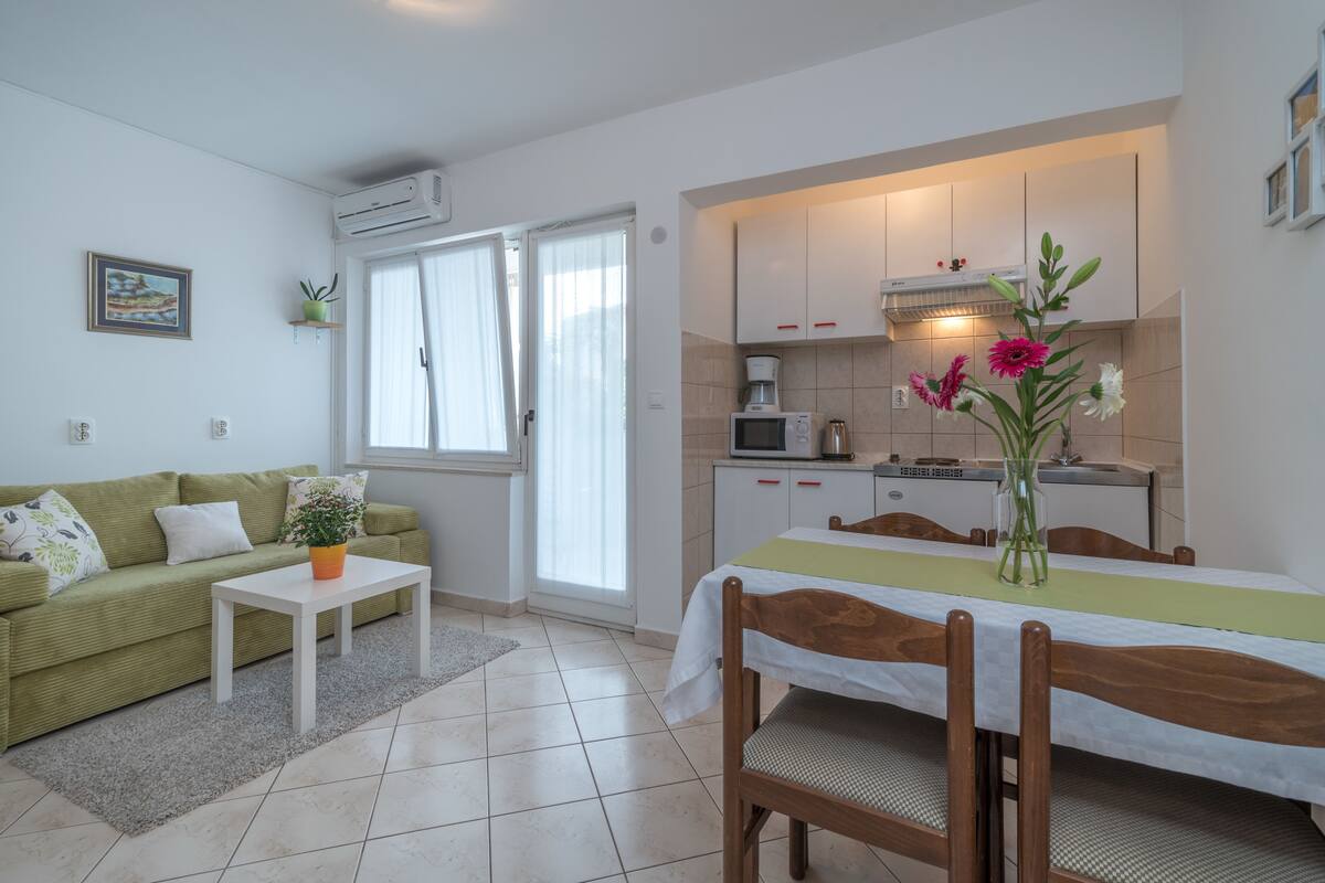 A compact living space with a green sofa, a small table, a dining table with chairs, a kitchen area with cabinets and appliances, and a window with curtains.