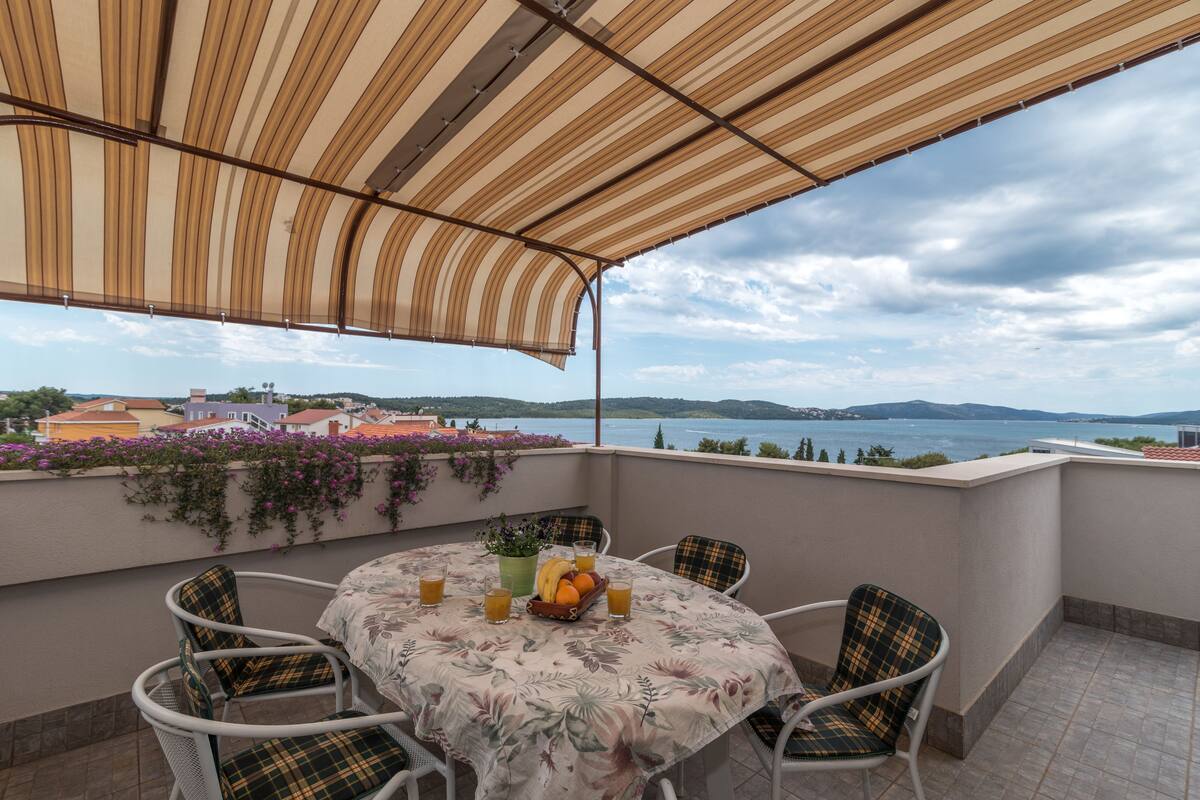 A terrace with a table set for two, a view of the sea, and a building in the background.