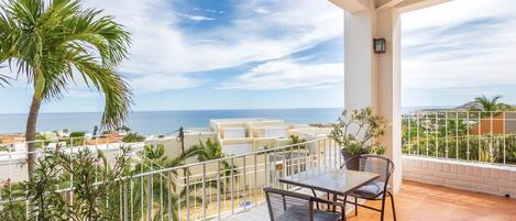 A balcony with a table and chairs, overlooking a coastal view with palm trees and buildings.