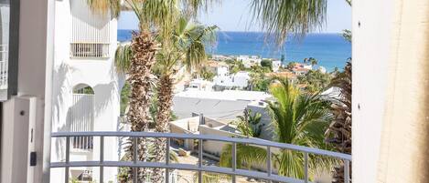 A balcony with a view of the sea, palm trees, and a white building.