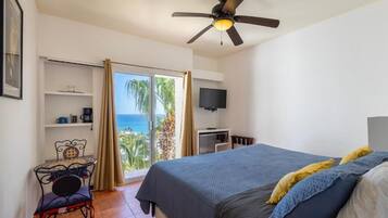 Une chambre avec un lit, un ventilateur de plafond, une télévision, un bureau et une vue sur l’océan, accessible par une porte coulissante.