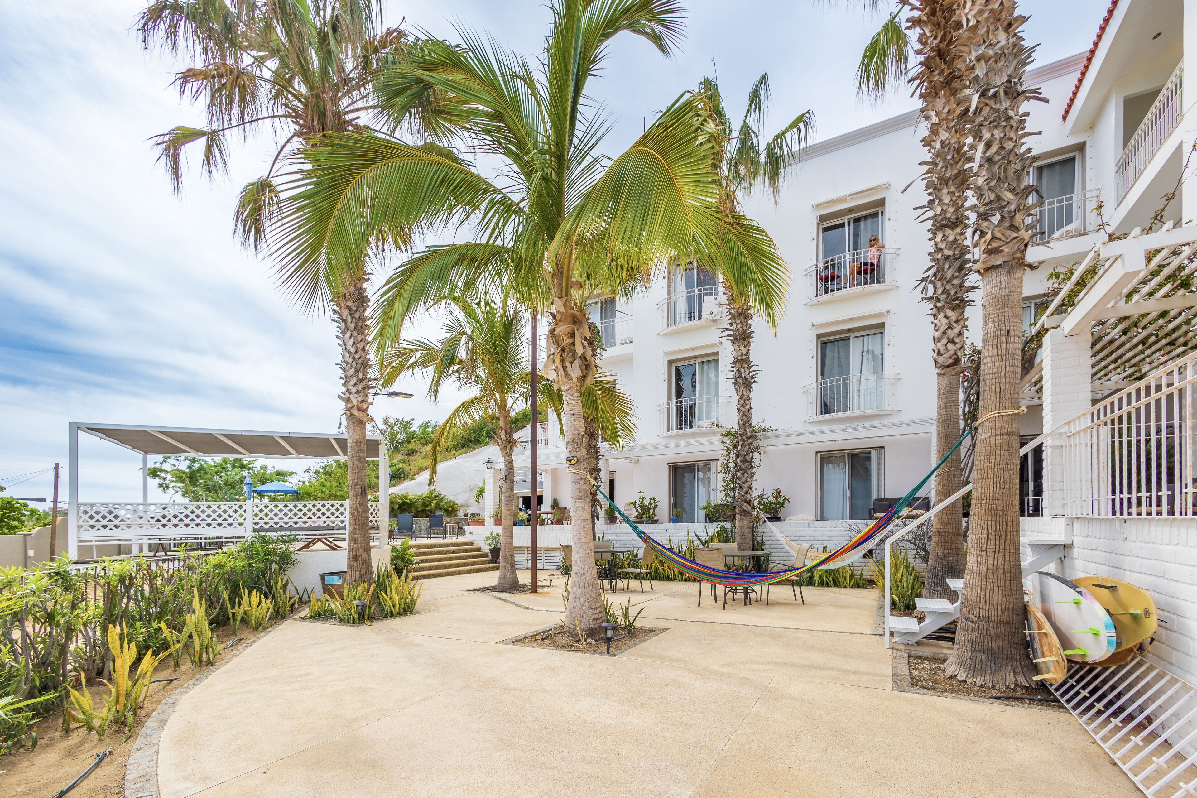 Une cour d’hôtel agrémentée de palmiers, d’un hamac et d’une planche de surf appuyée contre le bâtiment.