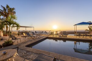 Zona de piscina con sillas de descanso y sombrillas, con vista a una playa al atardecer.