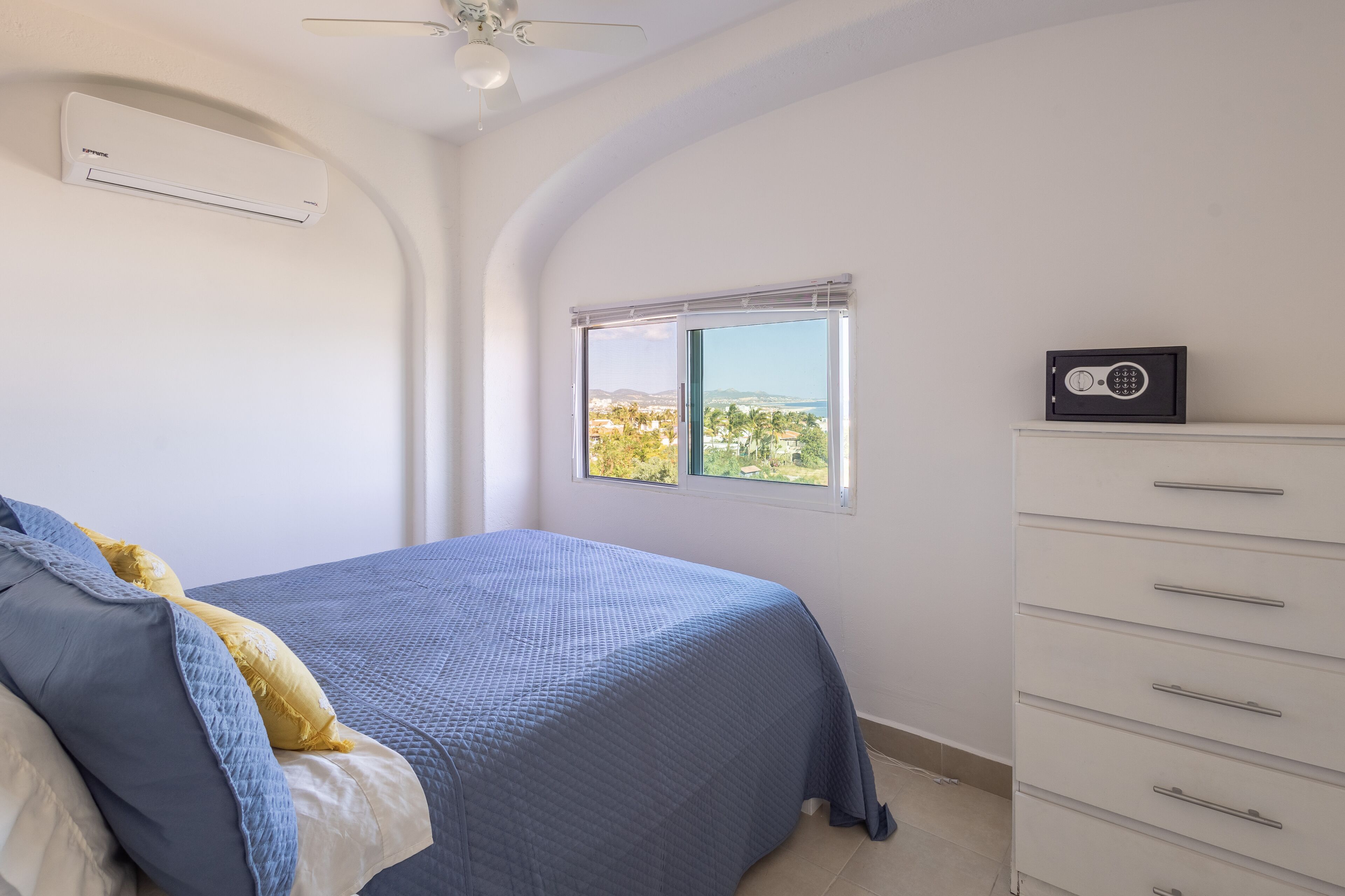Un dormitorio con una cama, un ventanal con vistas, un aparador blanco y un aire acondicionado.