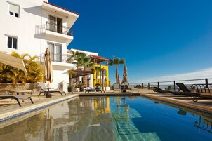 A swimming pool with lounge chairs and umbrellas, a white building with balconies, and palm trees.