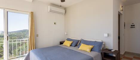 A bedroom with a large bed, a ceiling fan, an air conditioning unit, and a view of the outdoors through a sliding door.