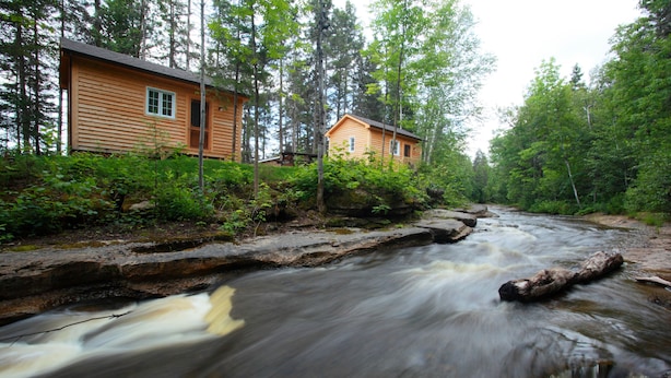 Une cabane aux murs en bois, entourée d’une forêt dense et bordée par une rivière aux eaux vives.